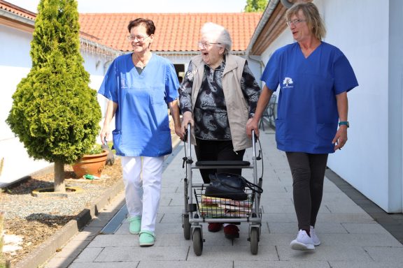 Zwei Pflegende begleiten eine ältere Frau mit einem Rollator auf einem Gehweg.