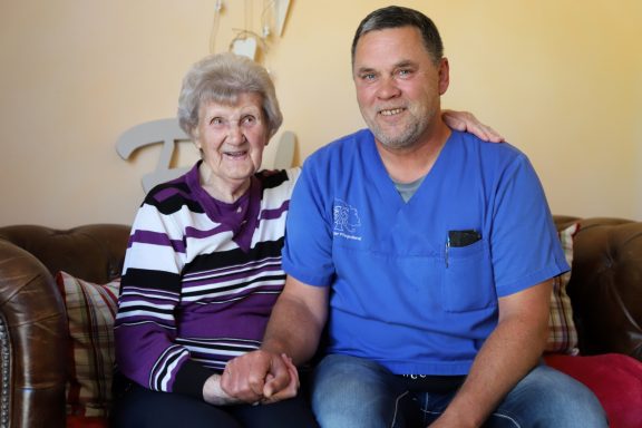 Ältere Frau und ein Mann in blauer Arbeitskleidung sitzen auf einer Couch und lächeln.