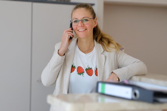 Lächelnde Frau mit Telefon, trägt ein T-Shirt mit Erdbeer-Motiv, am Schreibtisch.