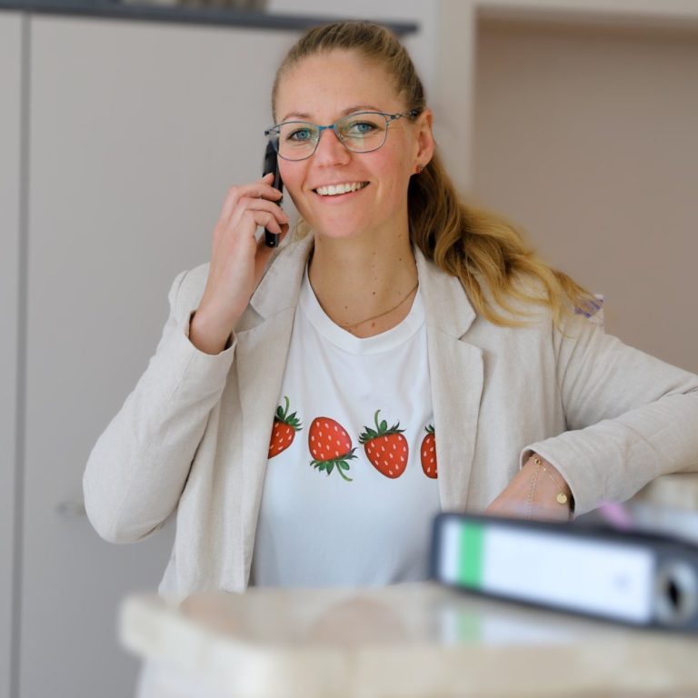 Frau mit blonden Haaren und Brille, am Telefon, trägt T-Shirt mit Erdbeeren.